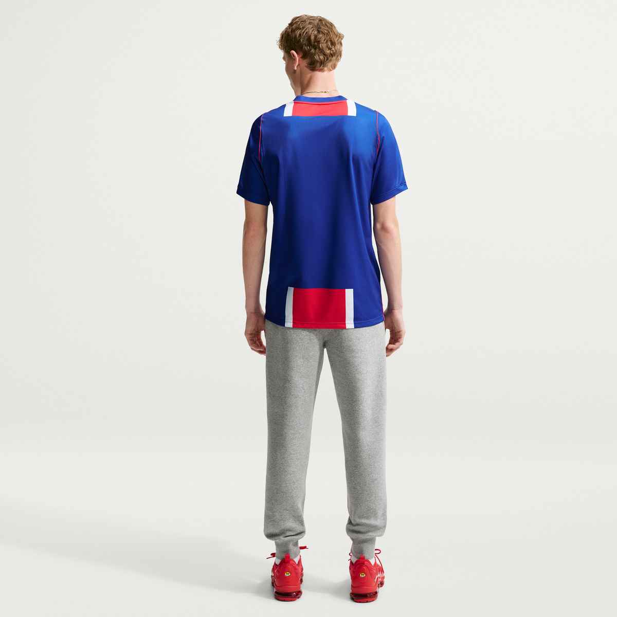 Person wearing a blue sports jersey with red and white accents, gray pants, and red shoes on a white background