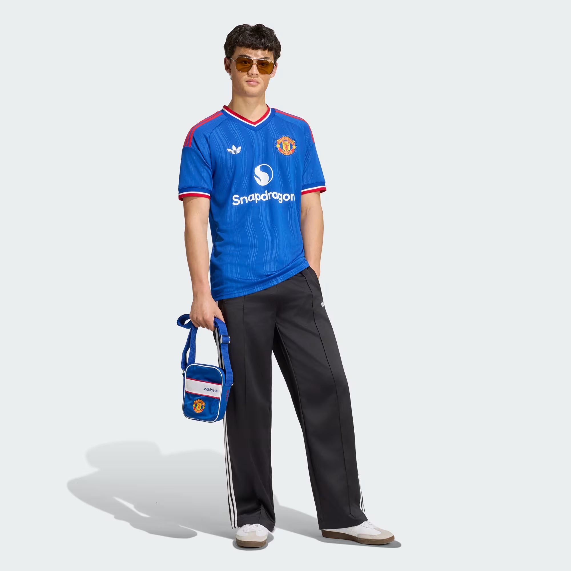 A model wearing a blue Manchester United soccer jersey with red and white trim, black track pants with white stripes, white sneakers, and sunglasses, holding a blue Manchester United shoulder bag against a light gray background.