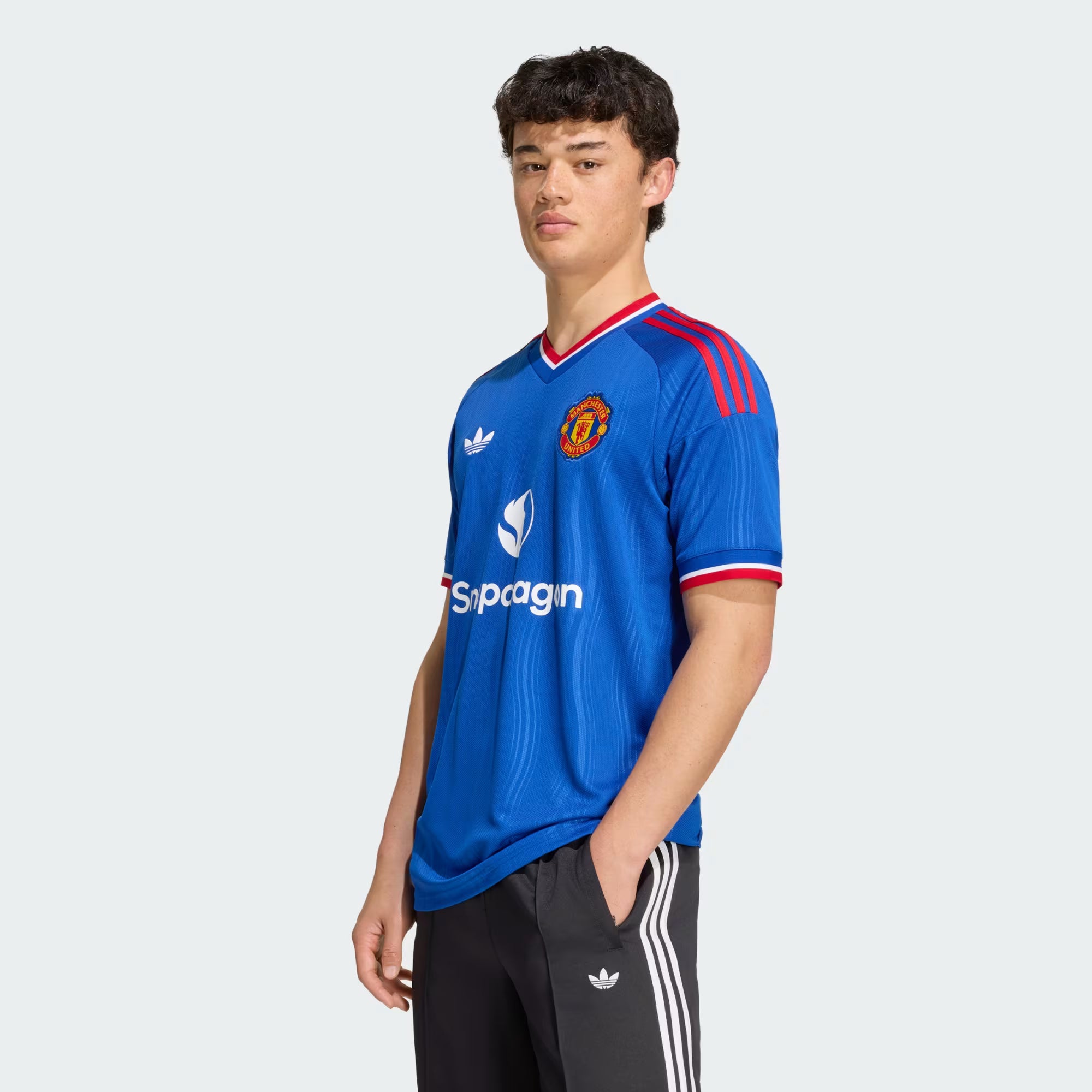 A young man modeling a blue Manchester United Adidas soccer jersey with red and white trim, paired with black Adidas track pants, against a light gray background.