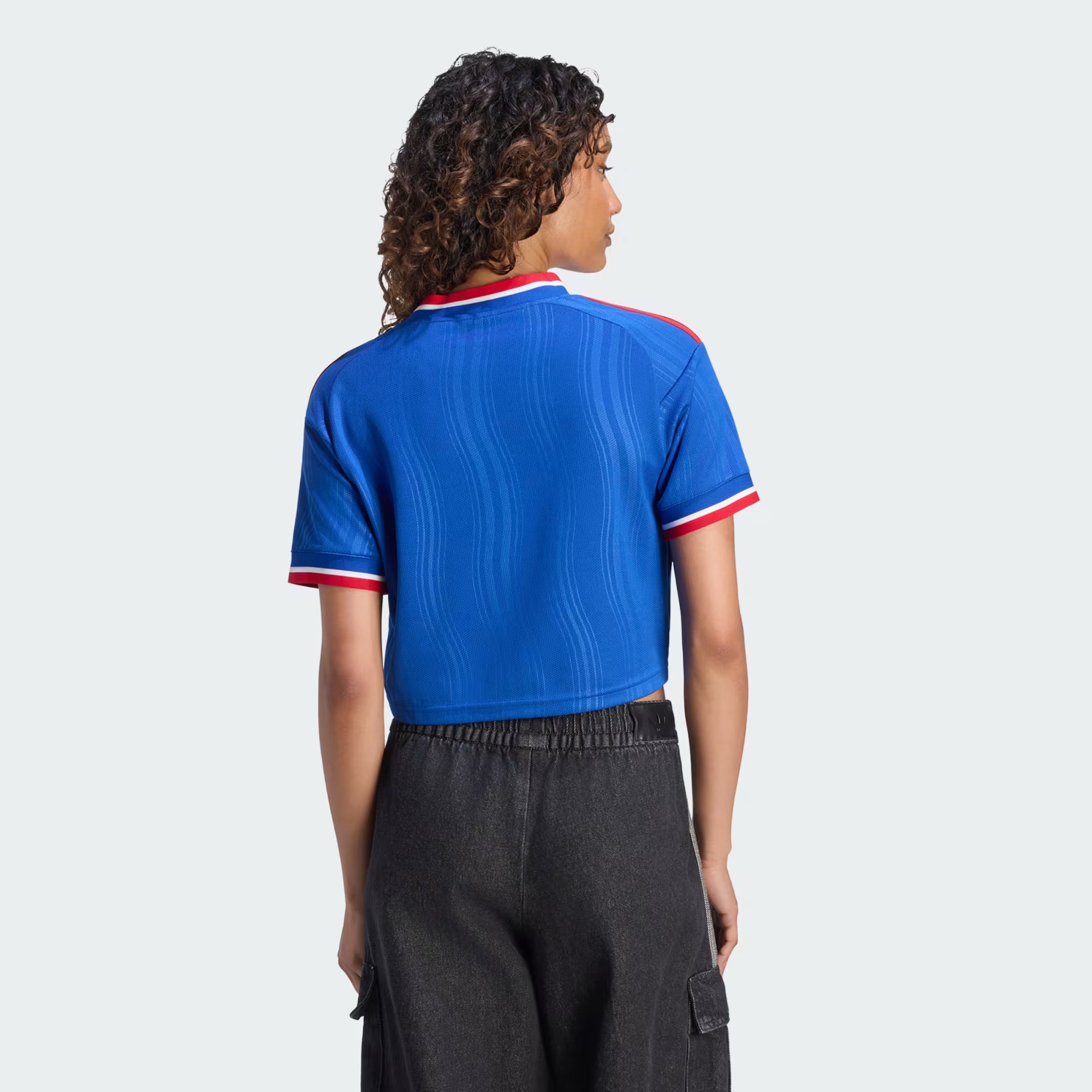 A model shown from the back wearing a blue short-sleeve cropped ribbed knit polo shirt with red and white trim on the collar and cuffs, paired with dark gray cargo pants, against a light gray background.