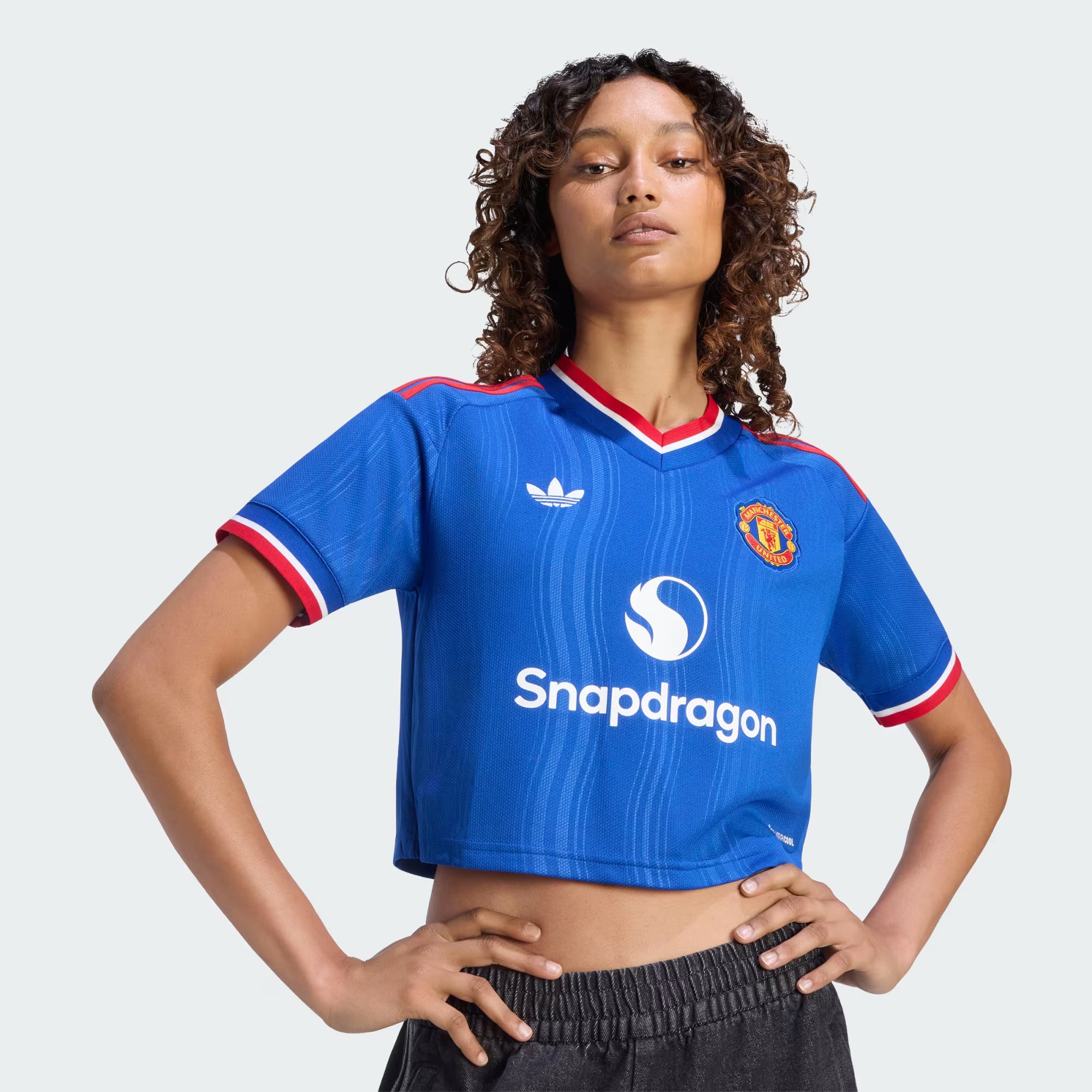 A model wearing a blue cropped short-sleeve Manchester United soccer jersey with red and white trim, featuring the Adidas logo, Snapdragon sponsor logo, and Manchester United crest.