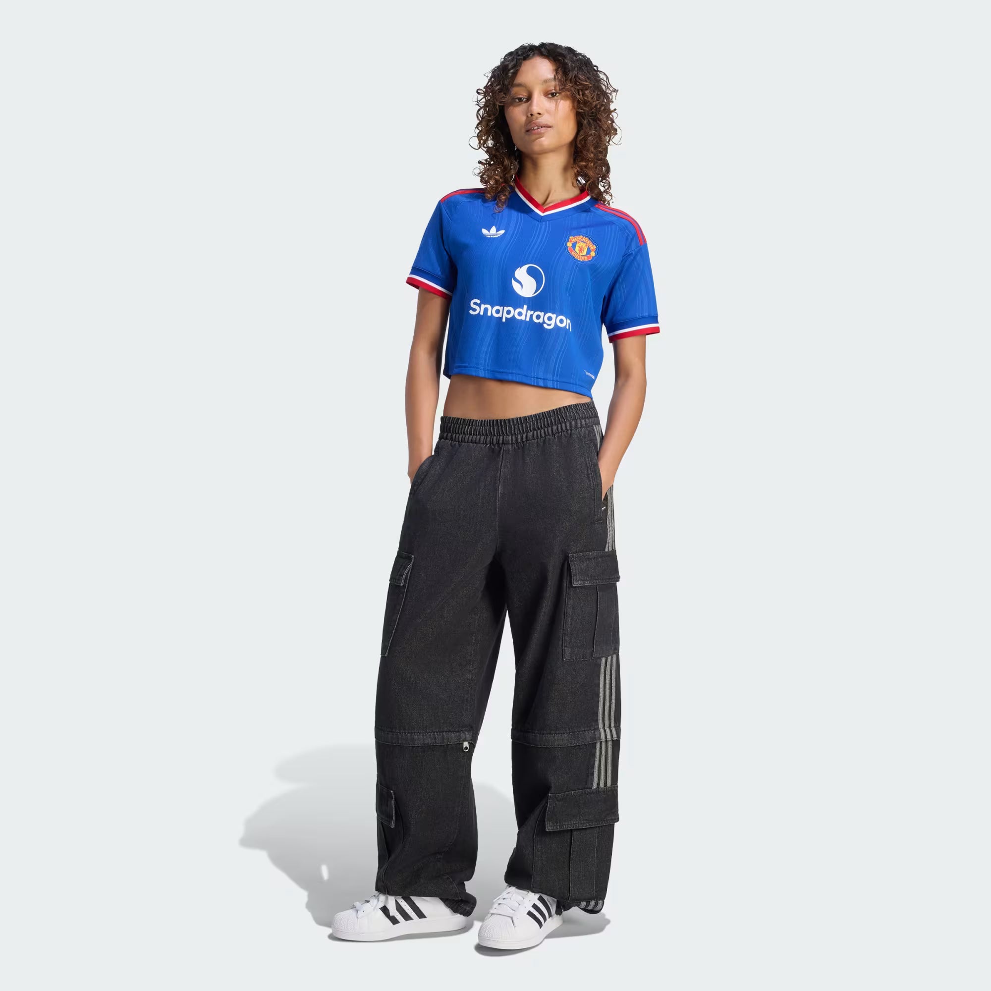 A model stands against a light gray studio background wearing a blue Manchester United cropped soccer jersey with white and red trim, paired with loose black cargo pants and white sneakers.