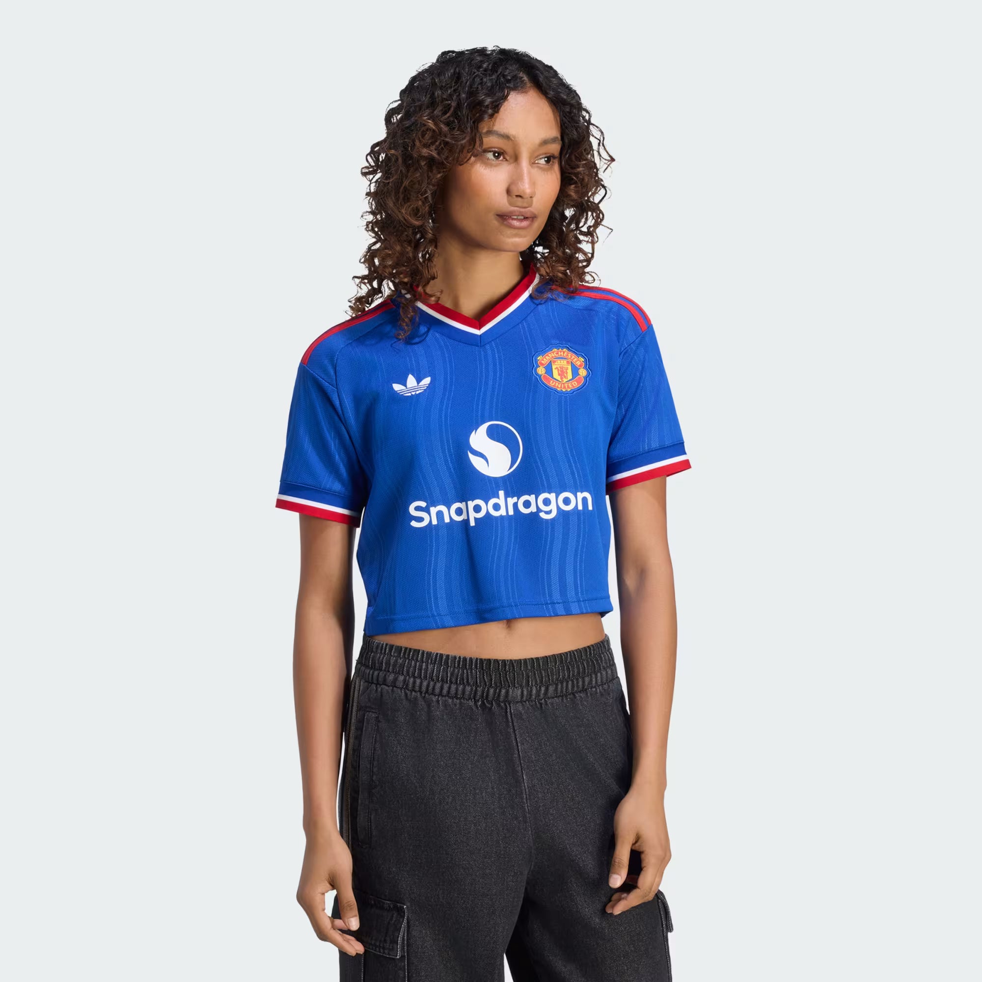 A model wearing a blue cropped Manchester United soccer jersey with red and white trim, featuring the Adidas logo, Manchester United crest, and Snapdragon sponsor, paired with dark gray cargo pants against a light gray background.