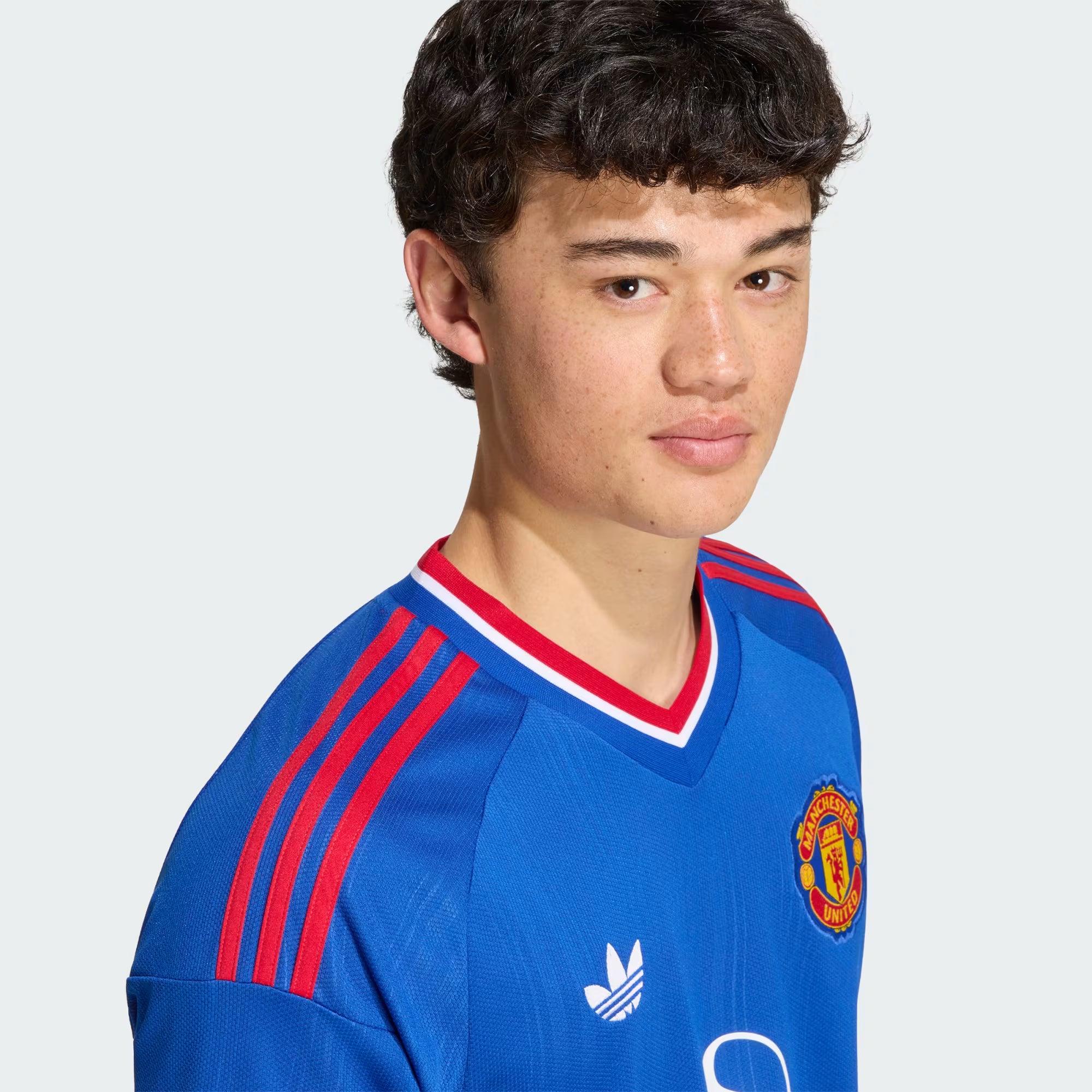 A young man wearing a blue Manchester United soccer jersey with red and white trim, featuring the Adidas logo and club crest, set against a light gray background.